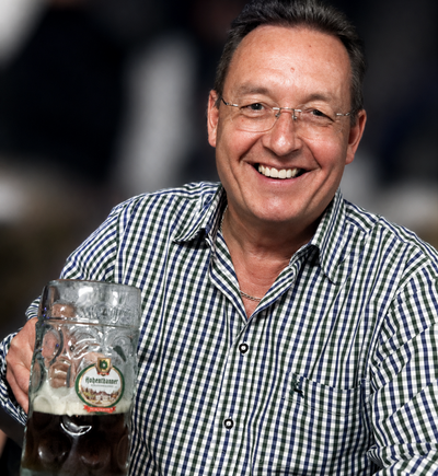 Markus Böckl hält lächelnd eine halbvolle Maß Bier in die Kamera; er trägt ein kariertes Hemd und sitzt am Tisch.